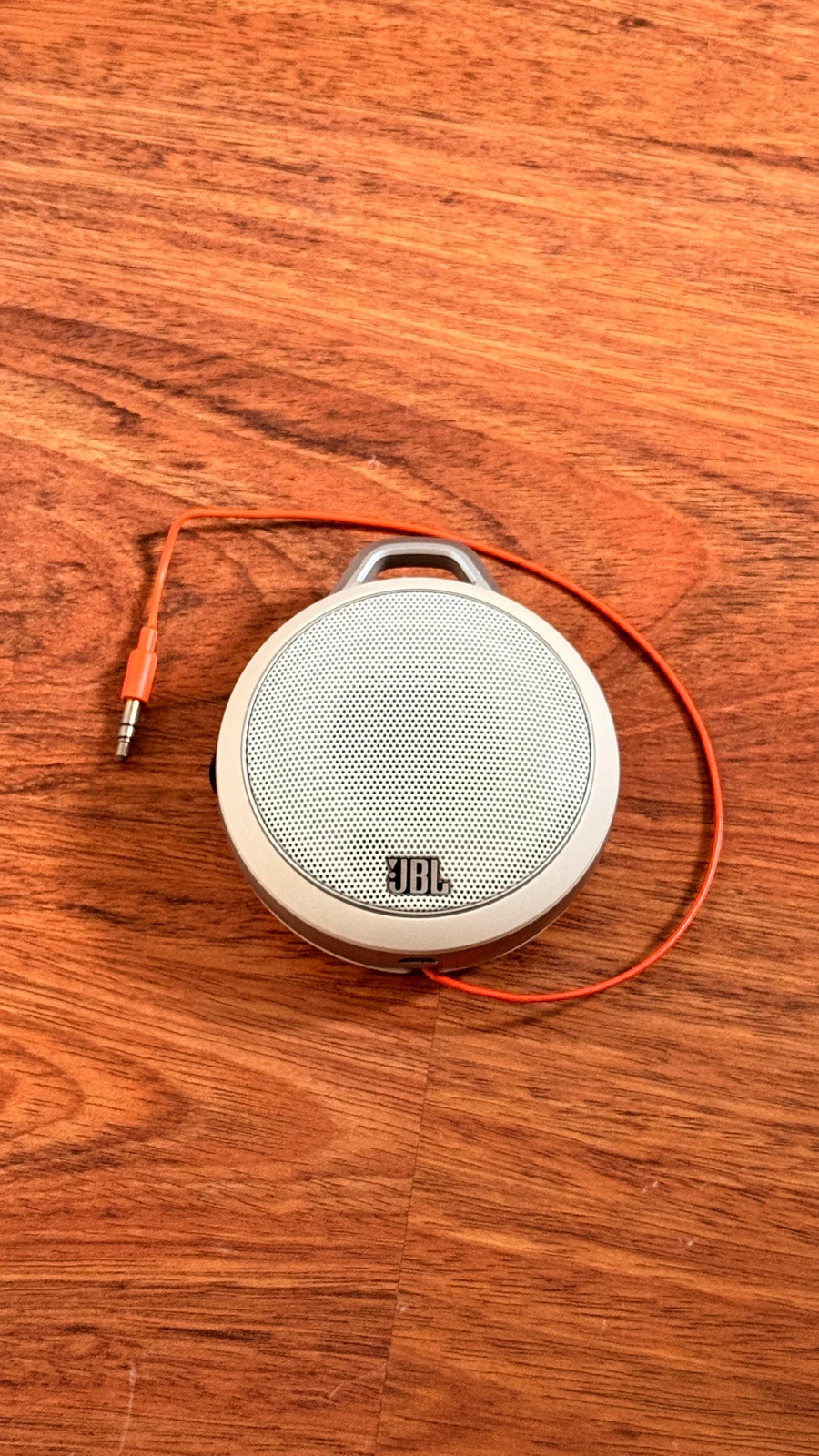 Ahşap yüzeyde ön yüzü görünen beyaz JBL hoparlör ve turuncu ses kablosu.