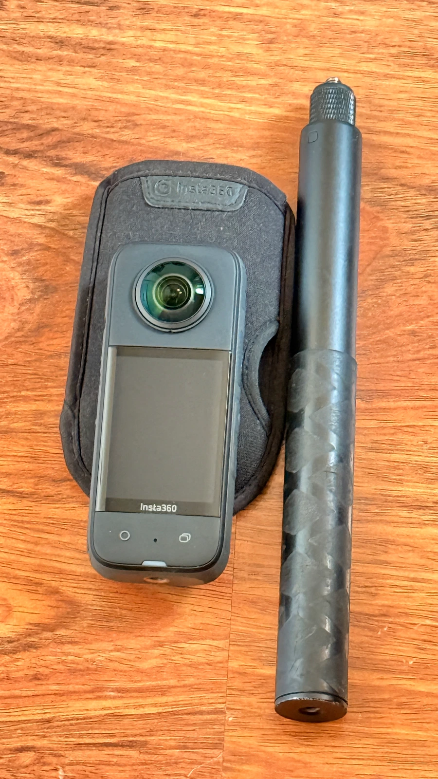 Ahşap yüzey üzerinde siyah Insta360 aksiyon kamerası, kılıfı ve uzun selfie çubuğu.